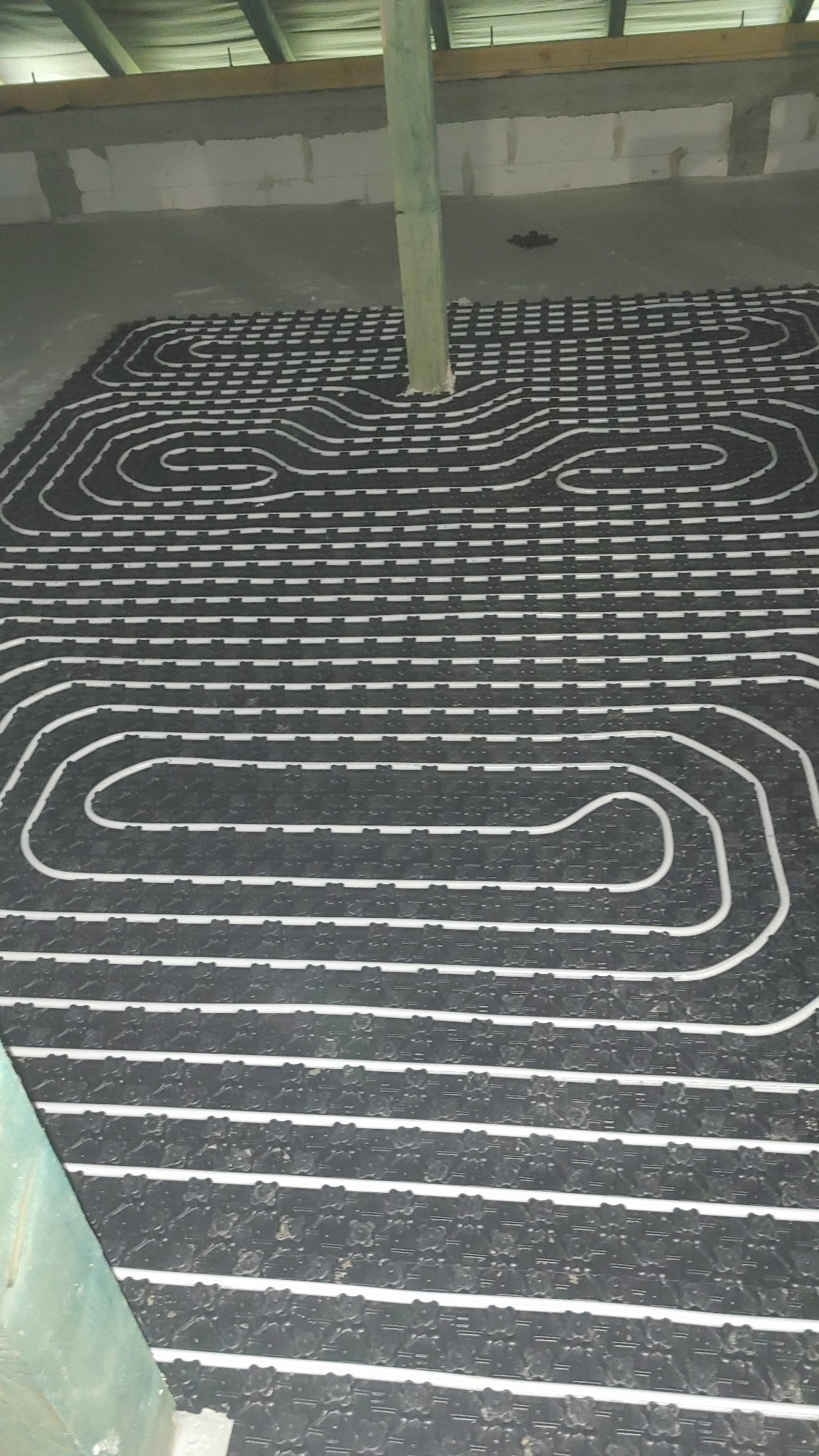 Rozłożona instalacja ogrzewania podłogowego na czarnej macie z widocznymi białymi rurkami, z drewnianym słupem przechodzącym przez instalację, w surowym stanie wykończenia.