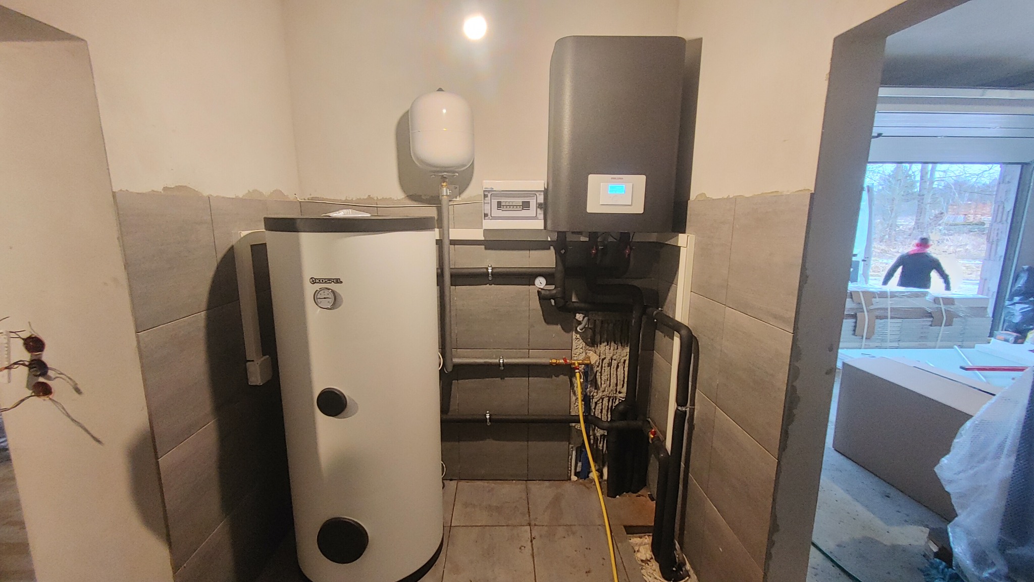 Bojler Kospel i szara pompa ciepła w kotłowni z szarymi płytkami, widoczne rury instalacyjne i otwarta przestrzeń za drzwiami.