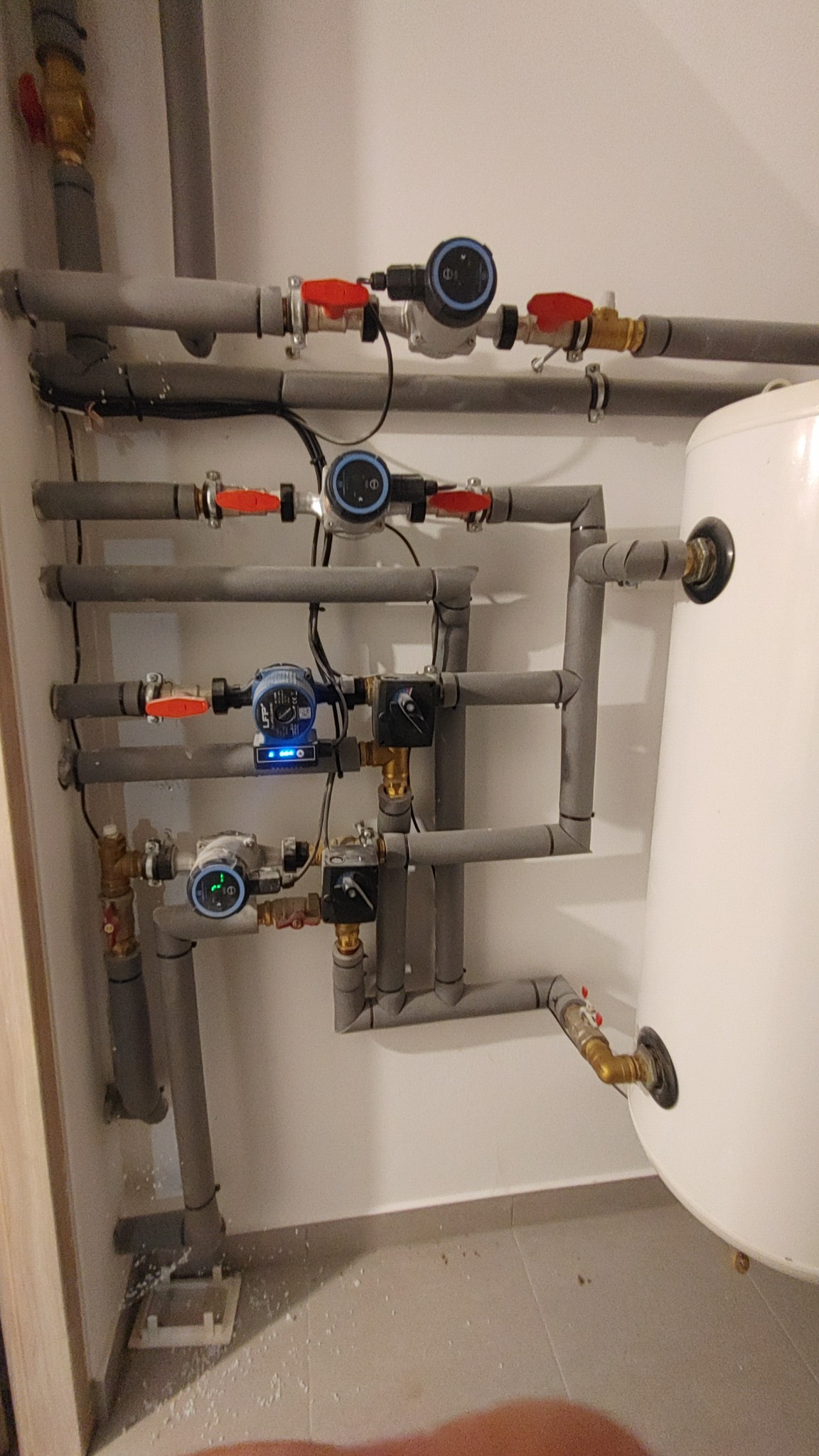 Rozbudowana instalacja hydrauliczna z rurami PCV, zaworami z czerwonymi rączkami i elektronicznymi pompami obiegowymi zamontowana na ścianie, obok biały zbiornik. Widoczne ślady po montażu...