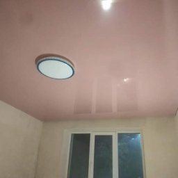 RemBudserwis - Błyszczący, różowy sufit napinany z zamontowanym okrągłym oświetleniem LED w pokoju z surowymi ścianami i oknem.