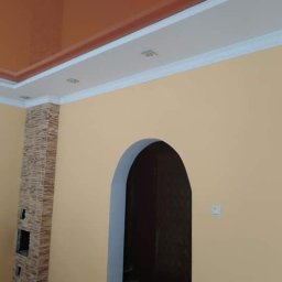 RemBudserwis - Wykończone wnętrze w Warszawie z łukowatym przejściem, beżowe ściany, błyszczący, pomarańczowy sufit podwieszany z białymi listwami i punktowym oświetleniem, oraz dekoracyjny filar z kamienia.