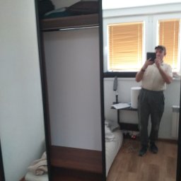 RemBudserwis - Wnętrze szafy z przesuwnymi drzwiami z lustrem, odbicie fotografa robiącego zdjęcie telefonem, widoczne półki wewnątrz i ubrania na górnej półce.