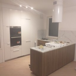 RemBudserwis - Nowoczesna kuchnia z białymi szafkami i wyspą z blatem imitującym marmur. Widoczne narzędzia i elementy montażowe na blacie wyspy. Minimalistyczny design.