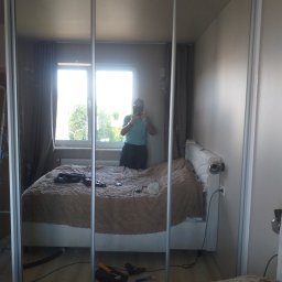 RemBudserwis - Szafa przesuwna z lustrem w sypialni w Warszawie. Widoczne odbicie osoby robiącej zdjęcie. Jasne wnętrze, łóżko z beżową narzutą i okno w tle.