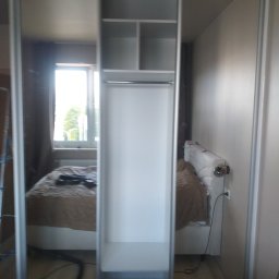 RemBudserwis - Wnętrze szafy wnękowej z przesuwnymi, lustrzanymi drzwiami, z widocznym fragmentem sypialni w odbiciu. Jasne półki i drążek na ubrania. Minimalistyczny design.