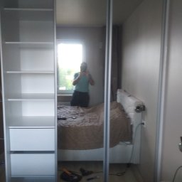 RemBudserwis - Biała szafa wnękowa z przesuwnymi, lustrzanymi drzwiami, odbijająca wnętrze sypialni z łóżkiem i oknem. Widoczne półki i szuflady, oraz osoba robiąca zdjęcie w lustrze.