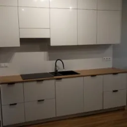 Biała, nowoczesna kuchnia z drewnianym blatem i czarną armaturą, oświetlona punktowymi lampami, prezentująca minimalistyczny design.