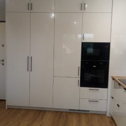 RemBudserwis - Biała, lakierowana szafa w zabudowie kuchennej z wbudowanym piekarnikiem i mikrofalówką, minimalistyczne uchwyty, drewniana podłoga, nowoczesne oświetlenie.