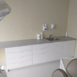 RemBudserwis - Białe szafki z szufladami i umywalką w gabinecie stomatologicznym. Blat w kolorze szarym. Na blacie dozowniki do mydła i ręczników. Krzesło dentystyczne w tle.