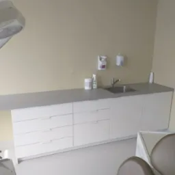 Białe szafki z szufladami i umywalką w gabinecie stomatologicznym. Blat w kolorze szarym. Na blacie dozowniki do mydła i ręczników. Krzesło dentystyczne w tle.