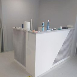 RemBudserwis - Nowoczesna, biała lada recepcyjna z pionowymi panelami i ukośnym wzorem, w minimalistycznym wnętrzu. Widoczne akcesoria montażowe i taśma ostrzegawcza.