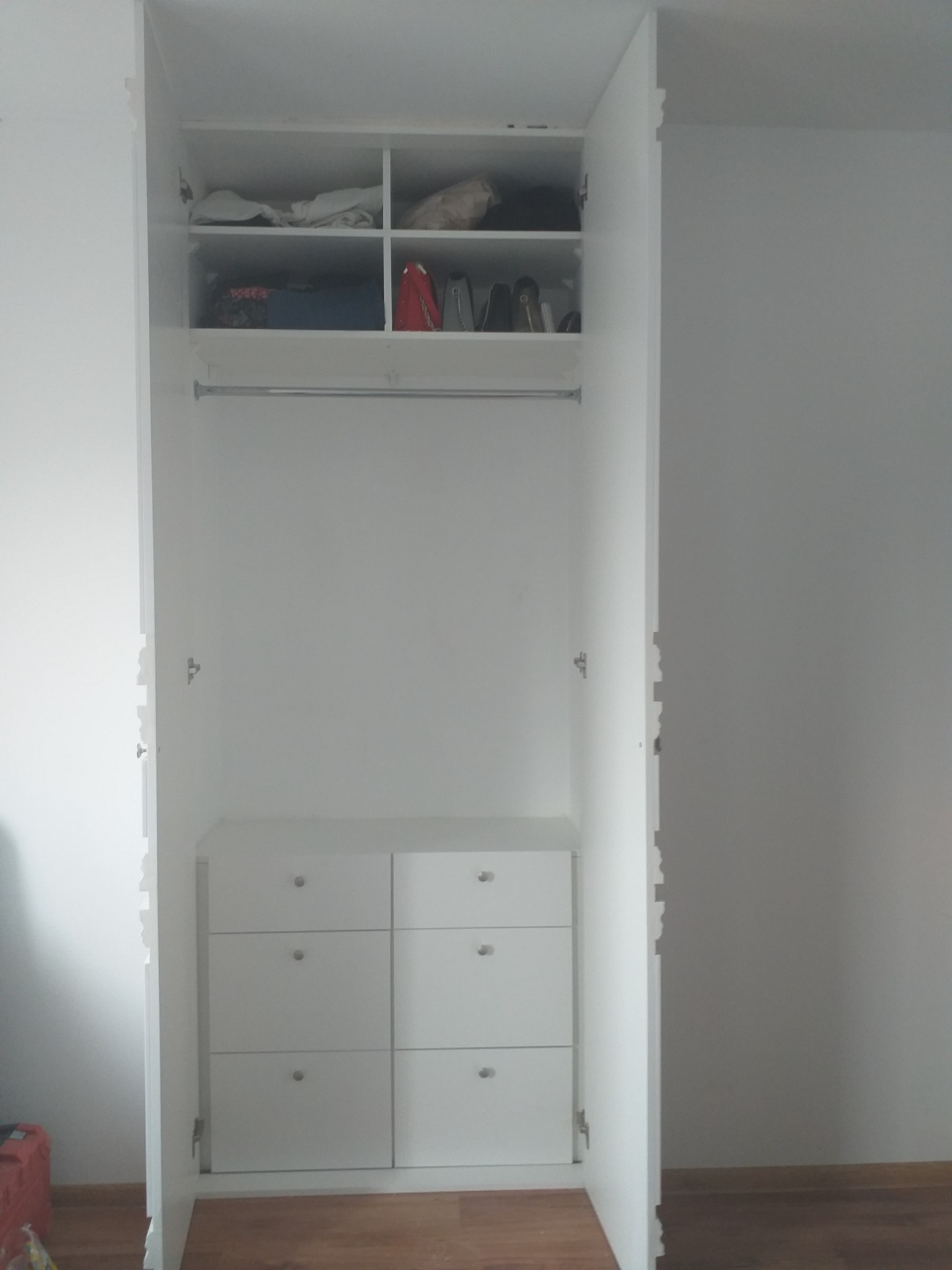 Wnętrze białej, otwartej szafy wnękowej z półkami u góry i sześcioma szufladami na dole, widoczne ubrania i akcesoria na półkach, drewniana podłoga.