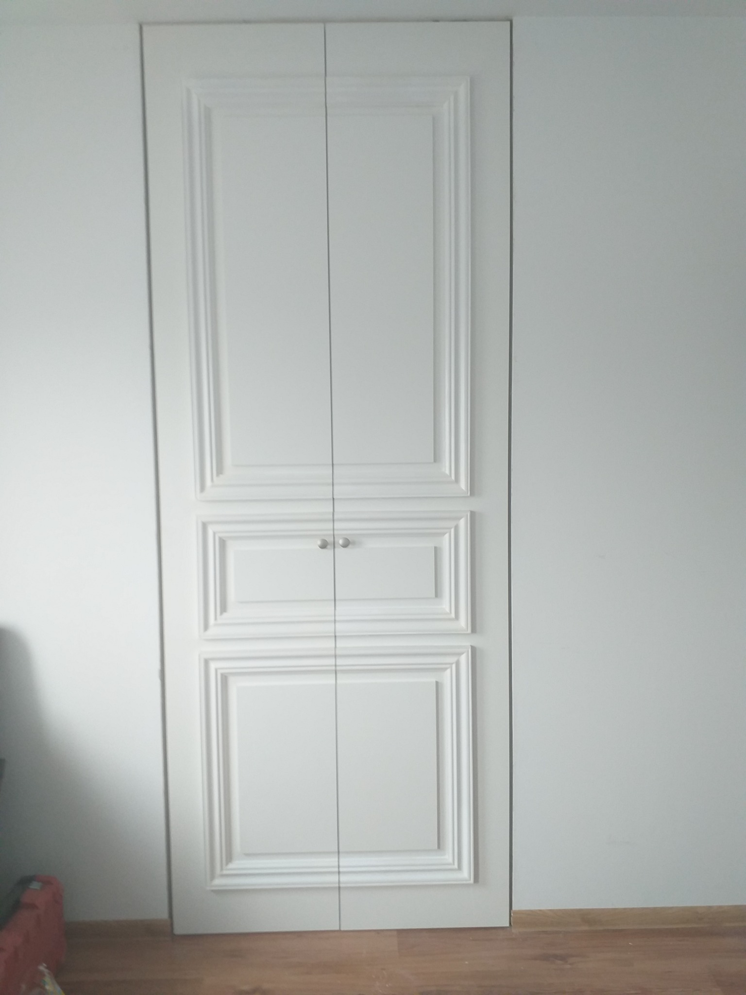 Białe, dwudrzwiowe, klasycznie zdobione drzwi wnękowe z gałkami, wkomponowane w ścianę, widoczne z bliska.