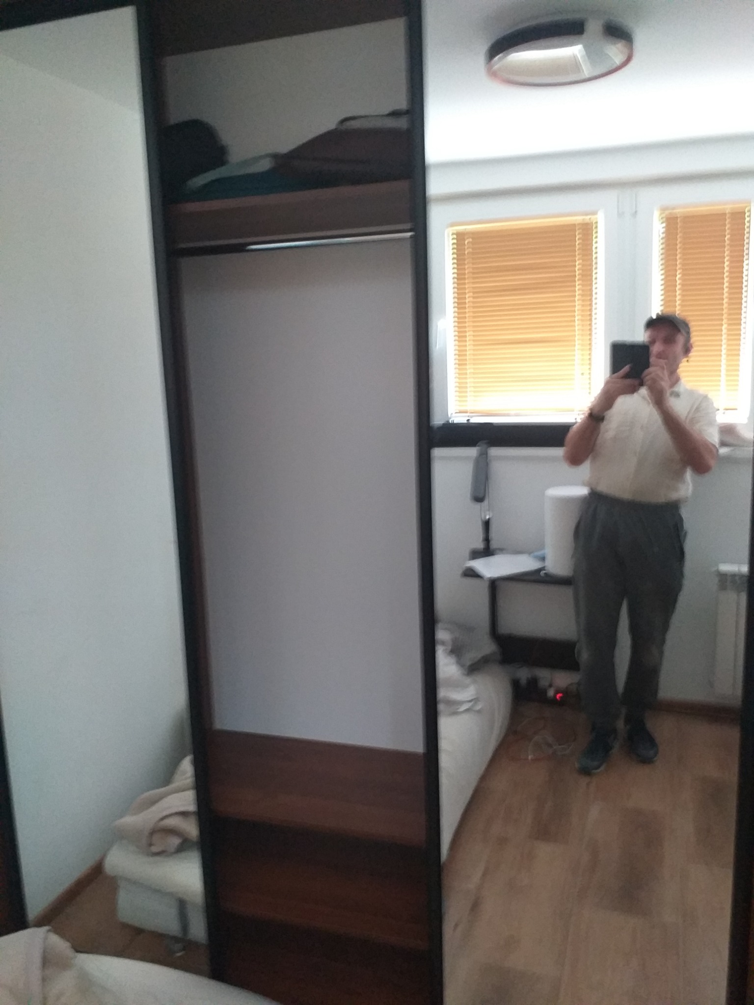 Wnętrze szafy z przesuwnymi drzwiami z lustrem, odbicie fotografa robiącego zdjęcie telefonem, widoczne półki wewnątrz i ubrania na górnej półce.