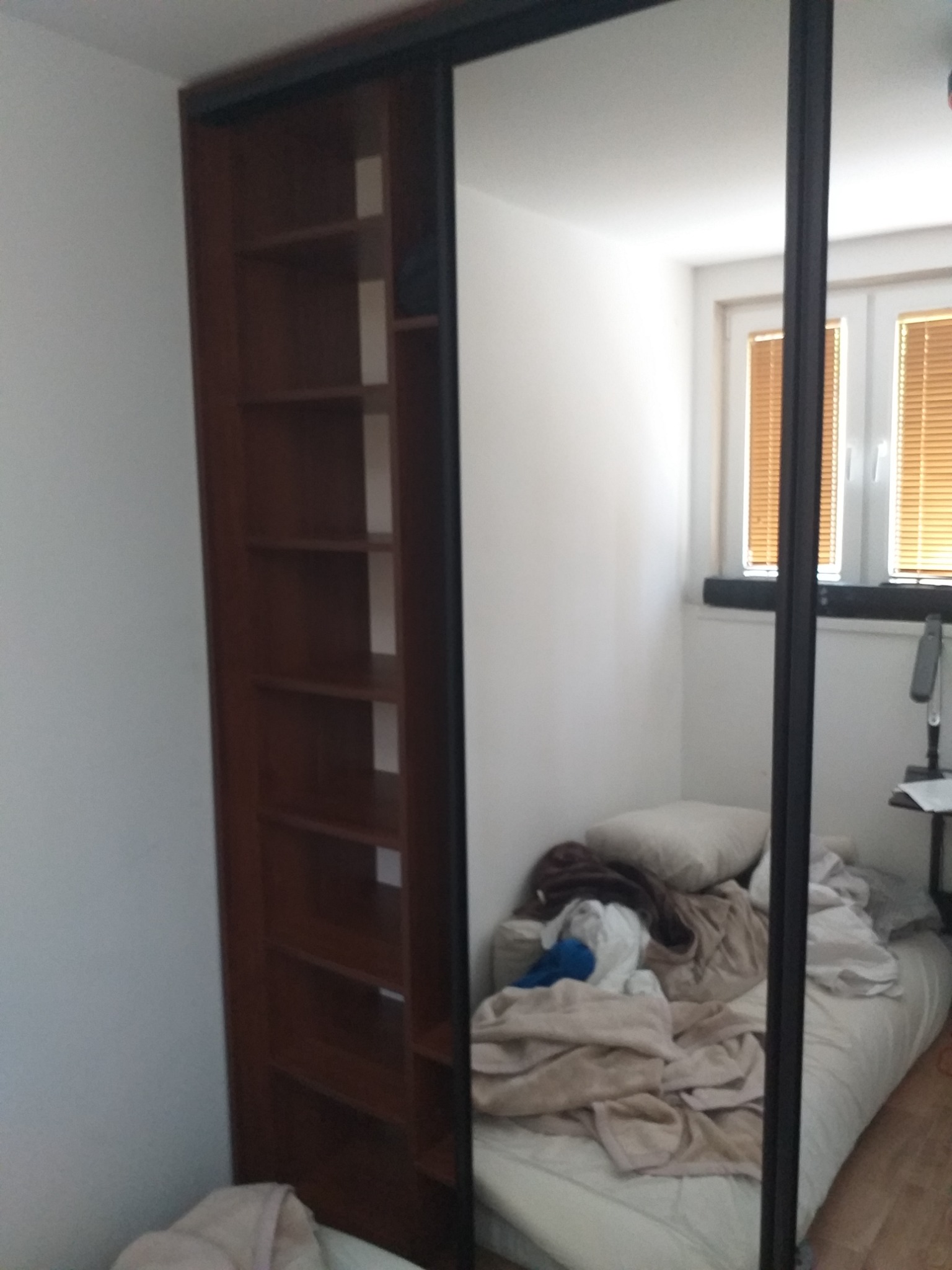 Wnętrze pokoju z szafą wnękową z przesuwnymi drzwiami lustrzanymi, widoczne półki w brązowej obudowie oraz odbicie łóżka z pościelą i okna.