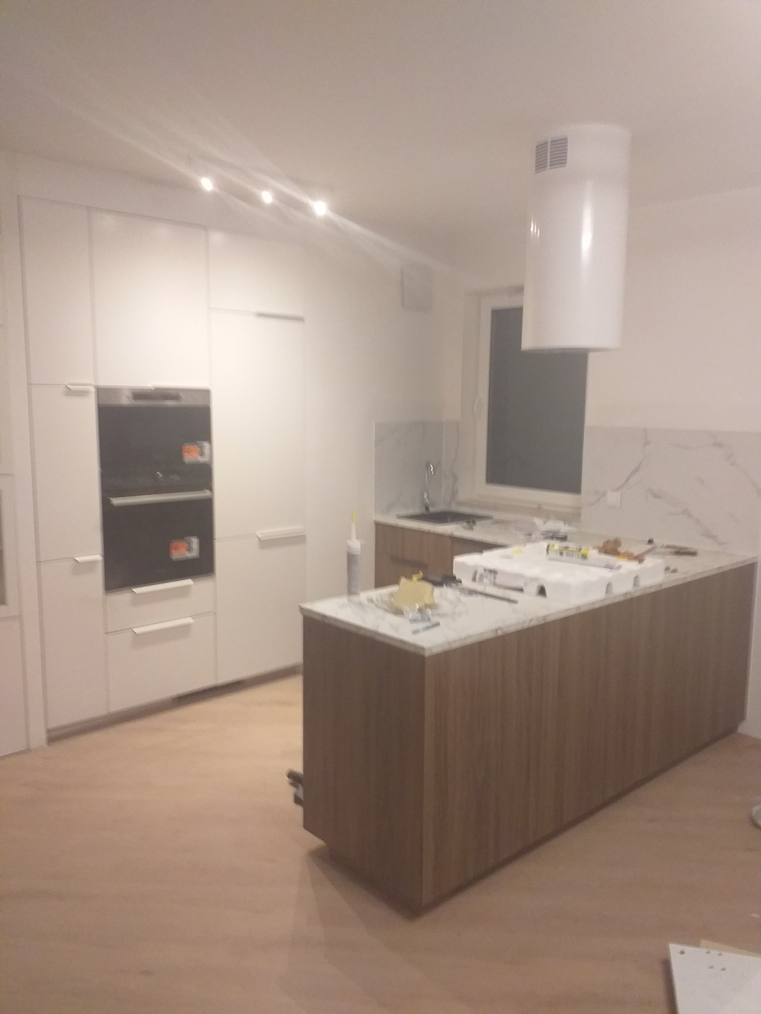 Nowoczesna kuchnia z białymi szafkami i wyspą z blatem imitującym marmur. Widoczne narzędzia i elementy montażowe na blacie wyspy. Minimalistyczny design.