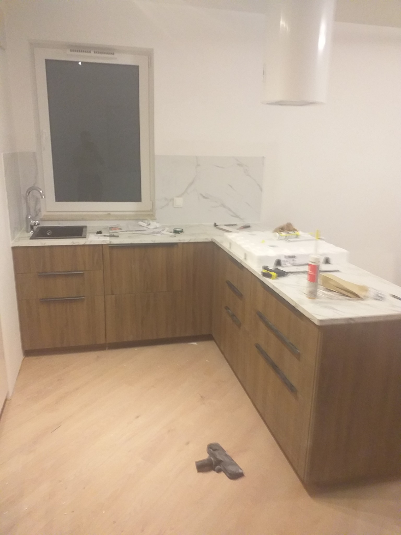 Kuchenna zabudowa z drewnianymi frontami i marmurowym blatem, zlewozmywak w oknie, okap. Częściowo ukończony montaż, widoczne narzędzia i materiały wykończeniowe.