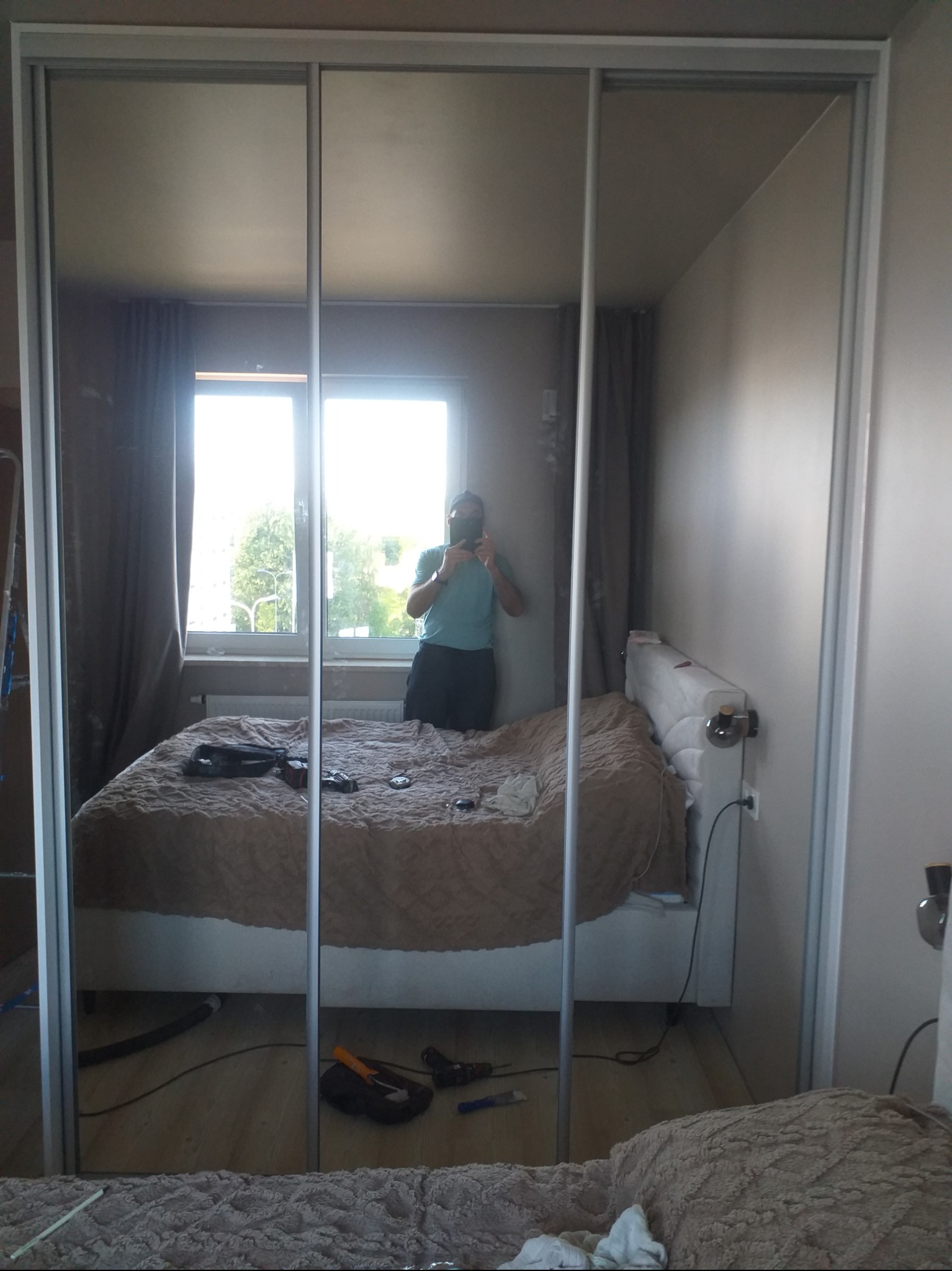 Szafa przesuwna z lustrem w sypialni w Warszawie. Widoczne odbicie osoby robiącej zdjęcie. Jasne wnętrze, łóżko z beżową narzutą i okno w tle.