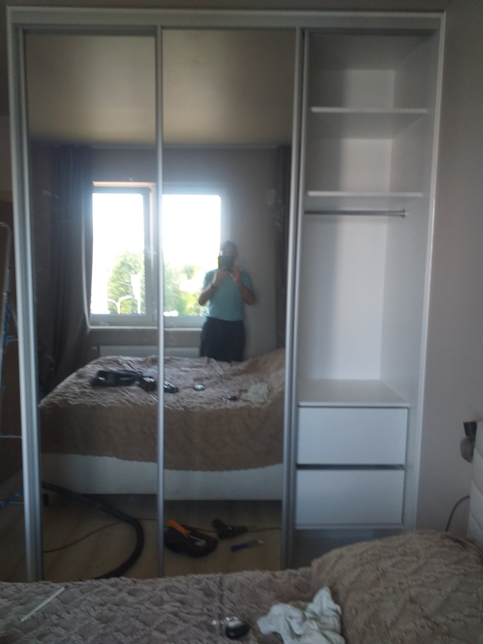 Biała szafa wnękowa z lustrzanymi drzwiami przesuwnymi i otwartymi półkami, widoczna w odbiciu sypialnia z oknem i osoba robiąca zdjęcie.