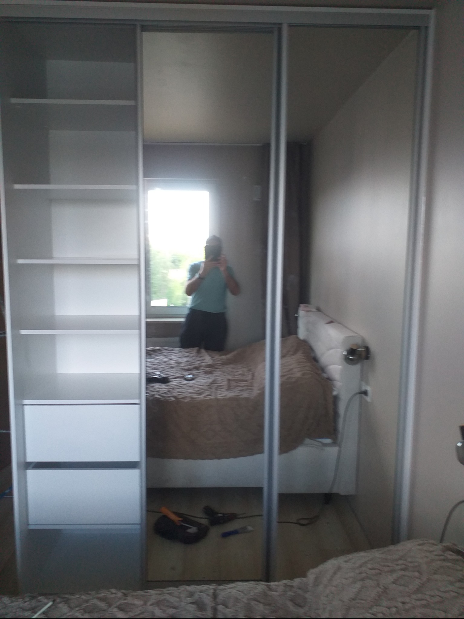 Biała szafa wnękowa z przesuwnymi, lustrzanymi drzwiami, odbijająca wnętrze sypialni z łóżkiem i oknem. Widoczne półki i szuflady, oraz osoba robiąca zdjęcie w lustrze.