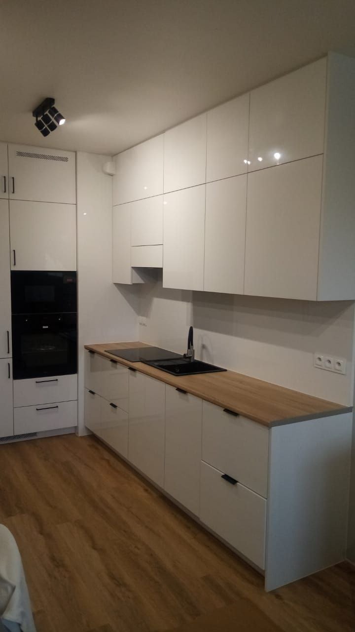 Biała, nowoczesna kuchnia z połyskiem, drewniany blat, czarny zlew i płyta grzewcza. Minimalistyczne uchwyty, oświetlenie punktowe, parkiet.