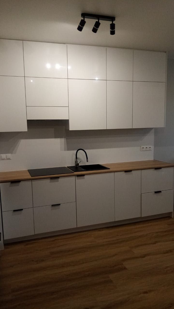 Biała, nowoczesna kuchnia z drewnianym blatem i czarną armaturą, oświetlona punktowymi lampami, prezentująca minimalistyczny design.