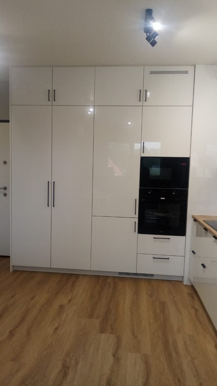 Biała, lakierowana szafa w zabudowie kuchennej z wbudowanym piekarnikiem i mikrofalówką, minimalistyczne uchwyty, drewniana podłoga, nowoczesne oświetlenie.