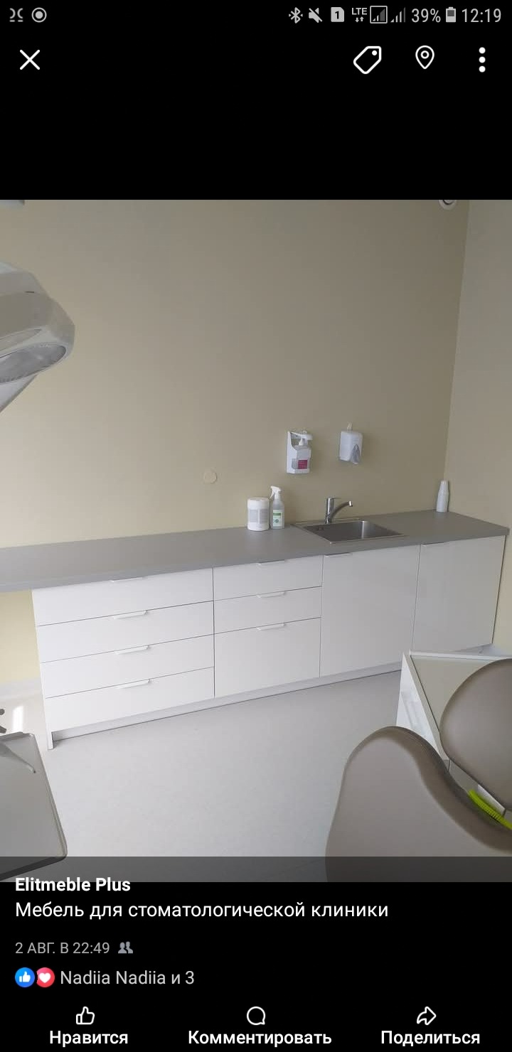 Białe szafki z szufladami i umywalką w gabinecie stomatologicznym. Blat w kolorze szarym. Na blacie dozowniki do mydła i ręczników. Krzesło dentystyczne w tle.