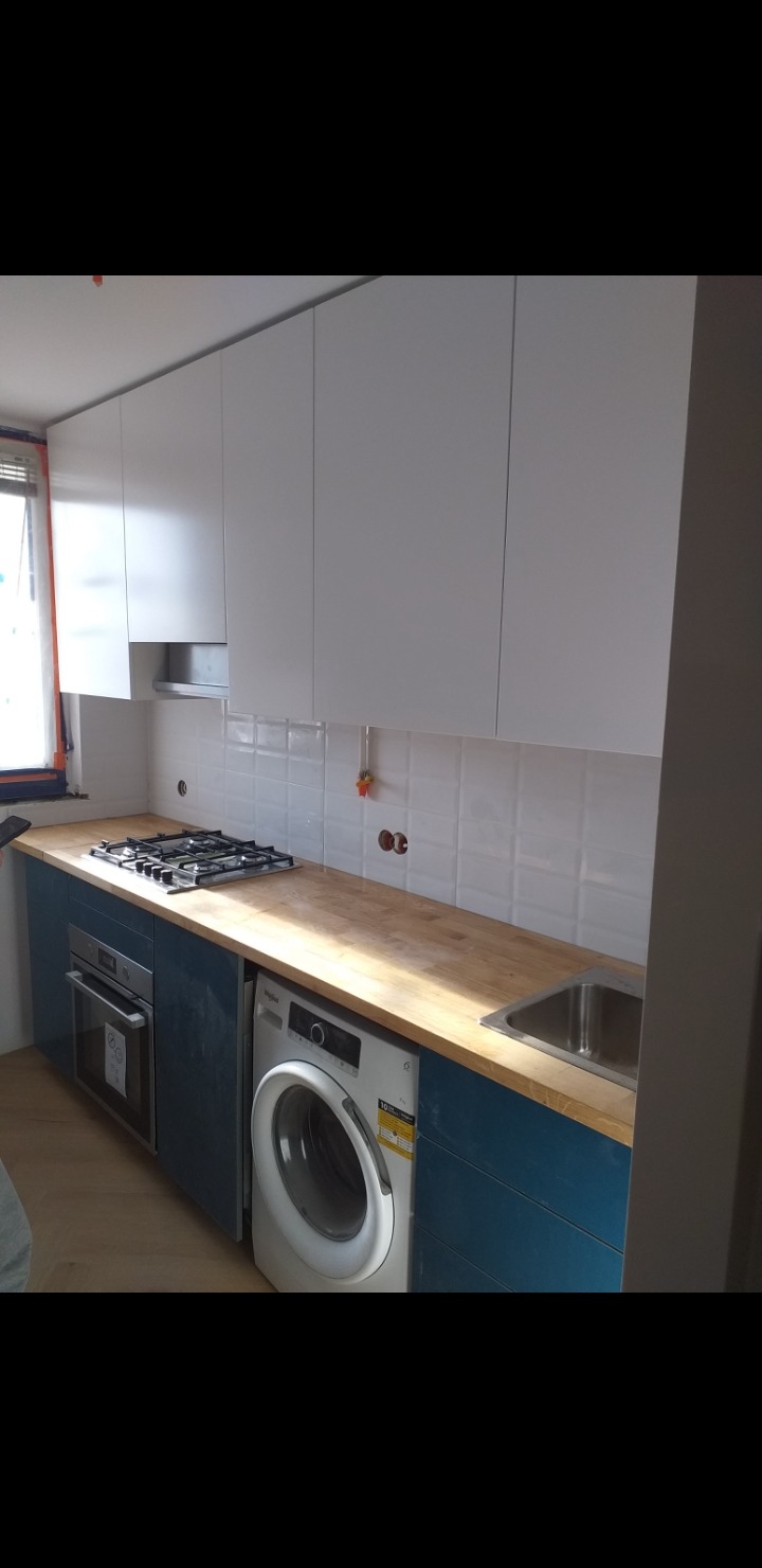 Nowoczesna kuchnia z białymi szafkami górnymi i niebieskimi dolnymi, drewnianym blatem, płytą gazową i pralką w zabudowie, wykończona białymi płytkami.