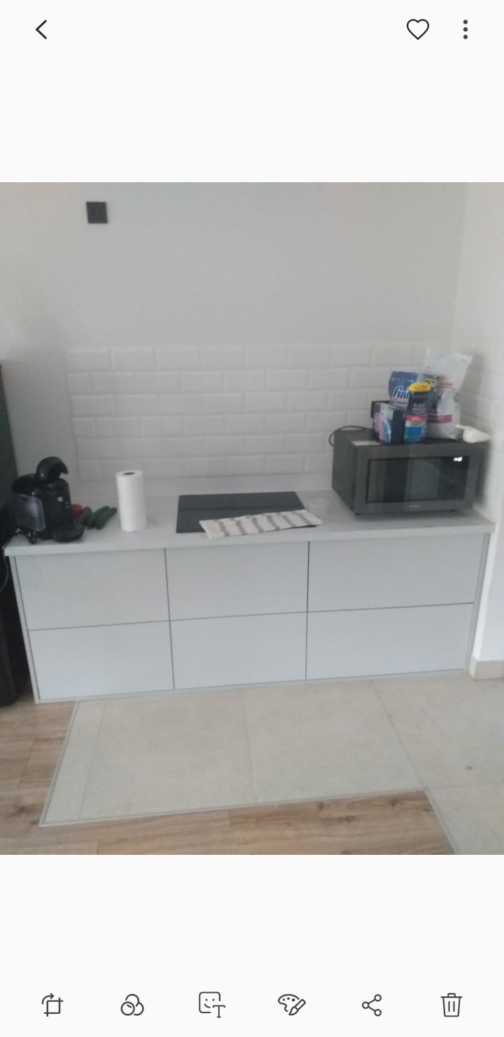 Minimalistyczna kuchnia: białe szafki dolne z blatem, płytki w stylu metro na ścianie, czarna płyta indukcyjna i mikrofalówka. Jasna podłoga z płytek.