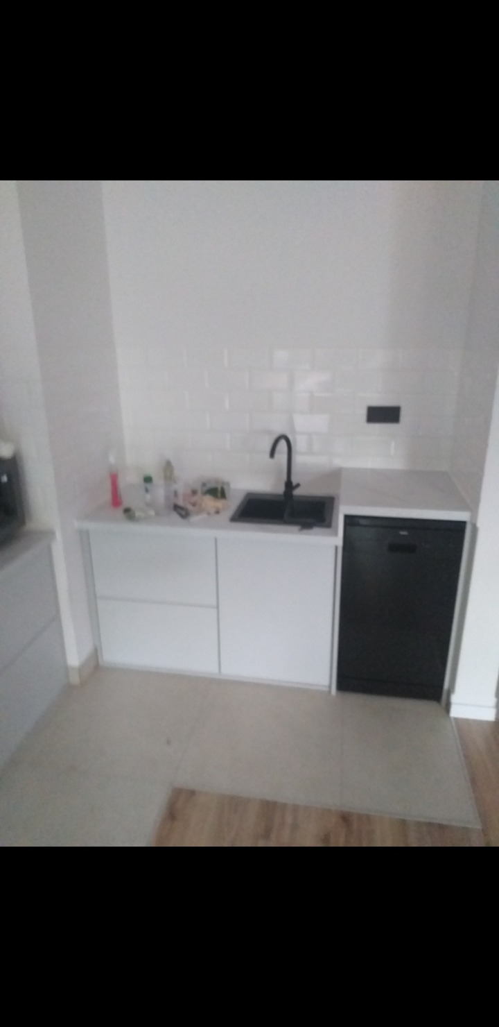 Biała zabudowa kuchenna z czarną armaturą i zmywarką. Minimalistyczny design z płytkami typu metro na ścianie. Ustawienie narożne, widok na fragment podłogi.