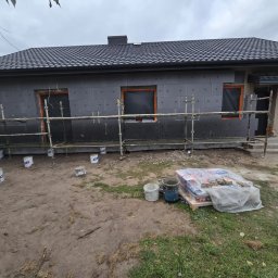 WARUSZEWSCY SPÓŁKA Z OGRANICZONĄ ODPOWIEDZIALNOŚCIĄ - Dom jednorodzinny w trakcie prac związanych z montażem izolacji termicznej elewacji, widoczne rusztowania, płyty izolacyjne oraz worki z klejem na ziemi.