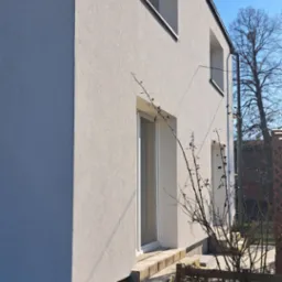 Wykończona elewacja domu jednorodzinnego z widocznym ociepleniem fundamentów, kamienne schody wejściowe i bezlistne krzewy na pierwszym planie, błękitne niebo w tle.