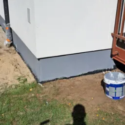 Dolna część elewacji budynku po malowaniu na szaro, widoczny kubeł z farbą i cień osoby malującej.