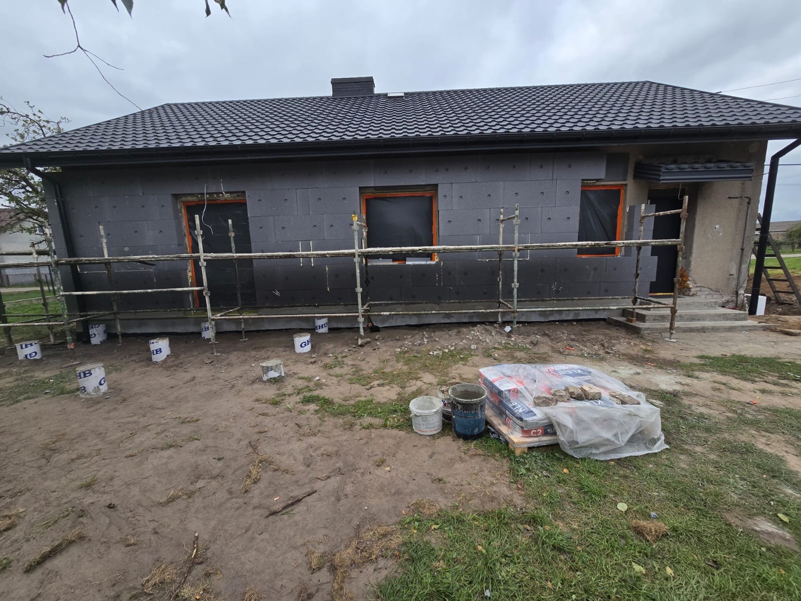 Dom jednorodzinny w trakcie prac związanych z montażem izolacji termicznej elewacji, widoczne rusztowania, płyty izolacyjne oraz worki z klejem na ziemi.