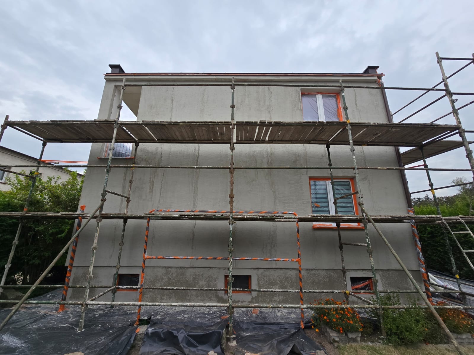 Dom w trakcie nakładania szarego tynku, widoczne rusztowanie z pomarańczowymi oznaczeniami, okna zabezpieczone folią, na dole rozłożona czarna folia ochronna.