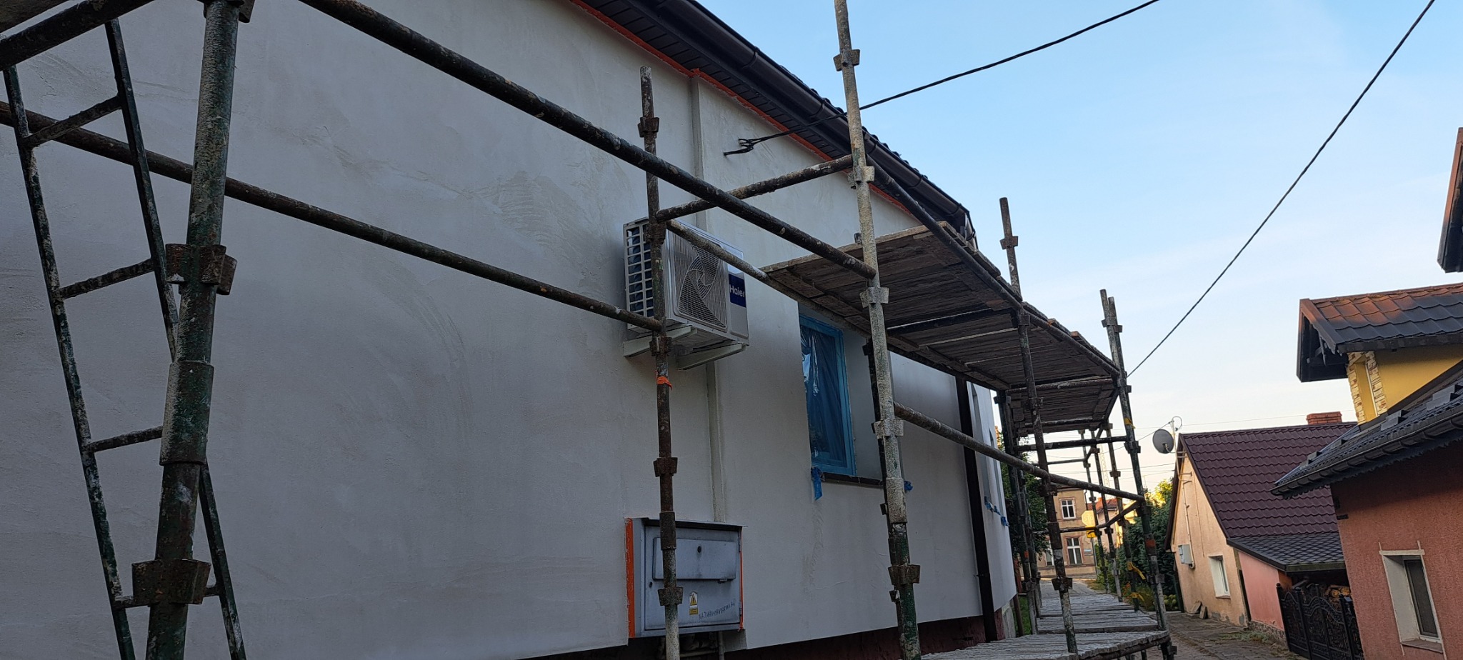 Świeżo otynkowana elewacja domu jednorodzinnego z klimatyzatorem, widoczna z poziomu rusztowania, okno zabezpieczone folią.