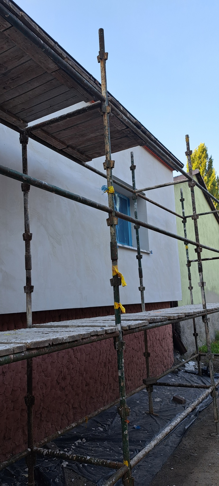 Świeżo otynkowana elewacja domu jednorodzinnego, widok z rusztowania, okno zabezpieczone folią, cokół z fakturą baranka w kolorze ceglastym, żółte taśmy zabezpieczające na rusztowaniu.