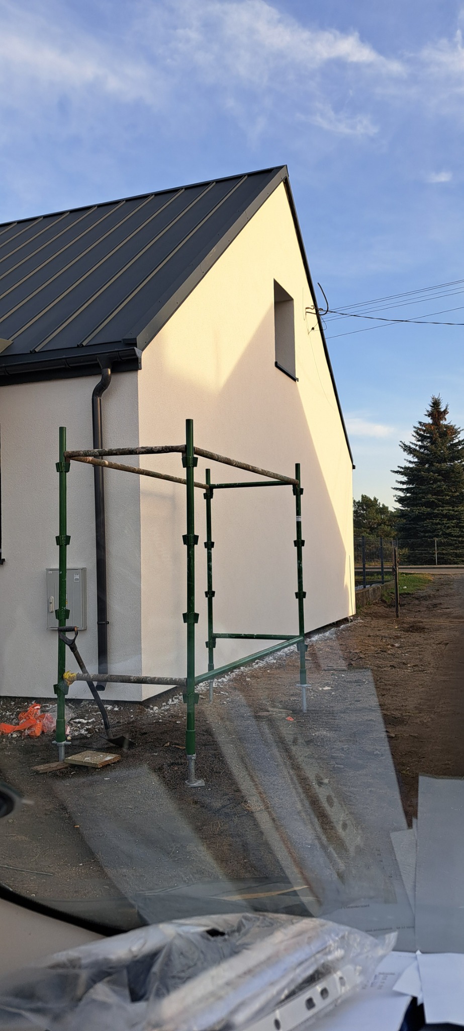 Fragment domu z jasną elewacją i ciemnym dachem, z zielonym rusztowaniem przy ścianie, widok zza szyby samochodu.