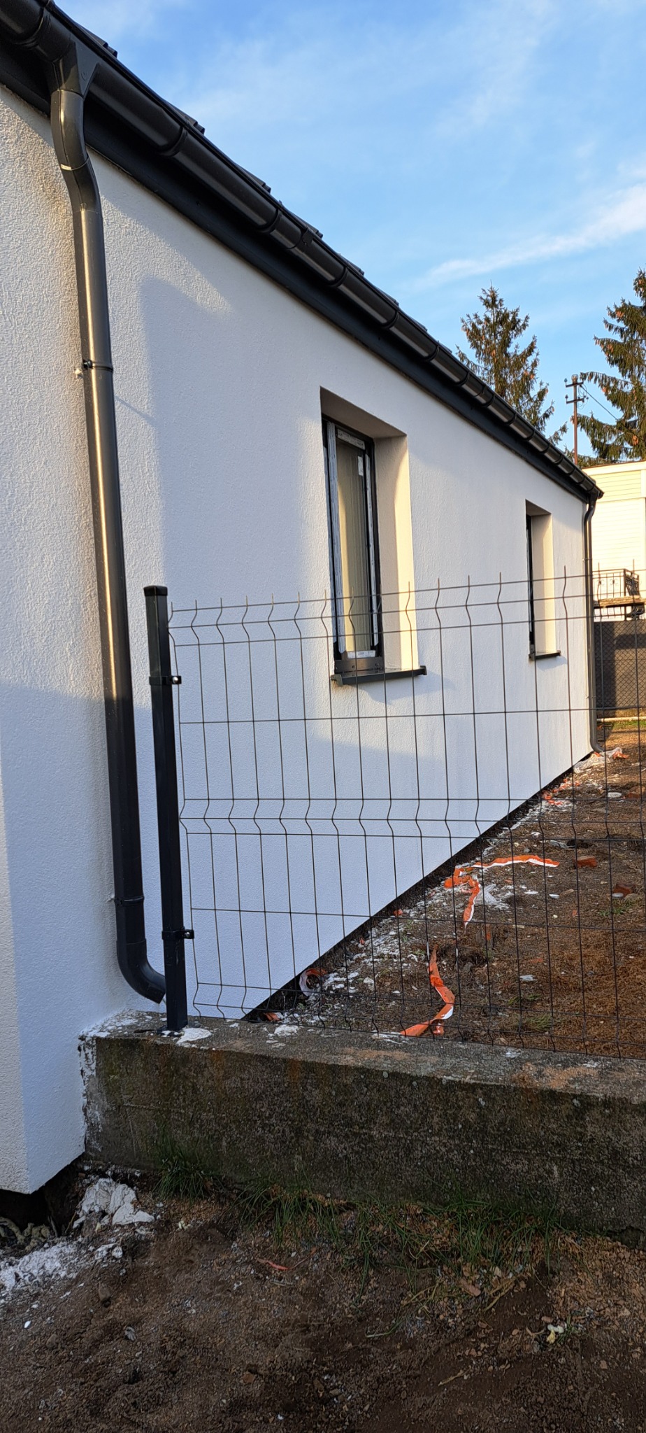 Świeżo otynkowana, biała ściana domu z czarnymi oknami i rynnami, widoczna zza metalowego ogrodzenia. Na ziemi resztki folii ochronnej i drobne gruz.
