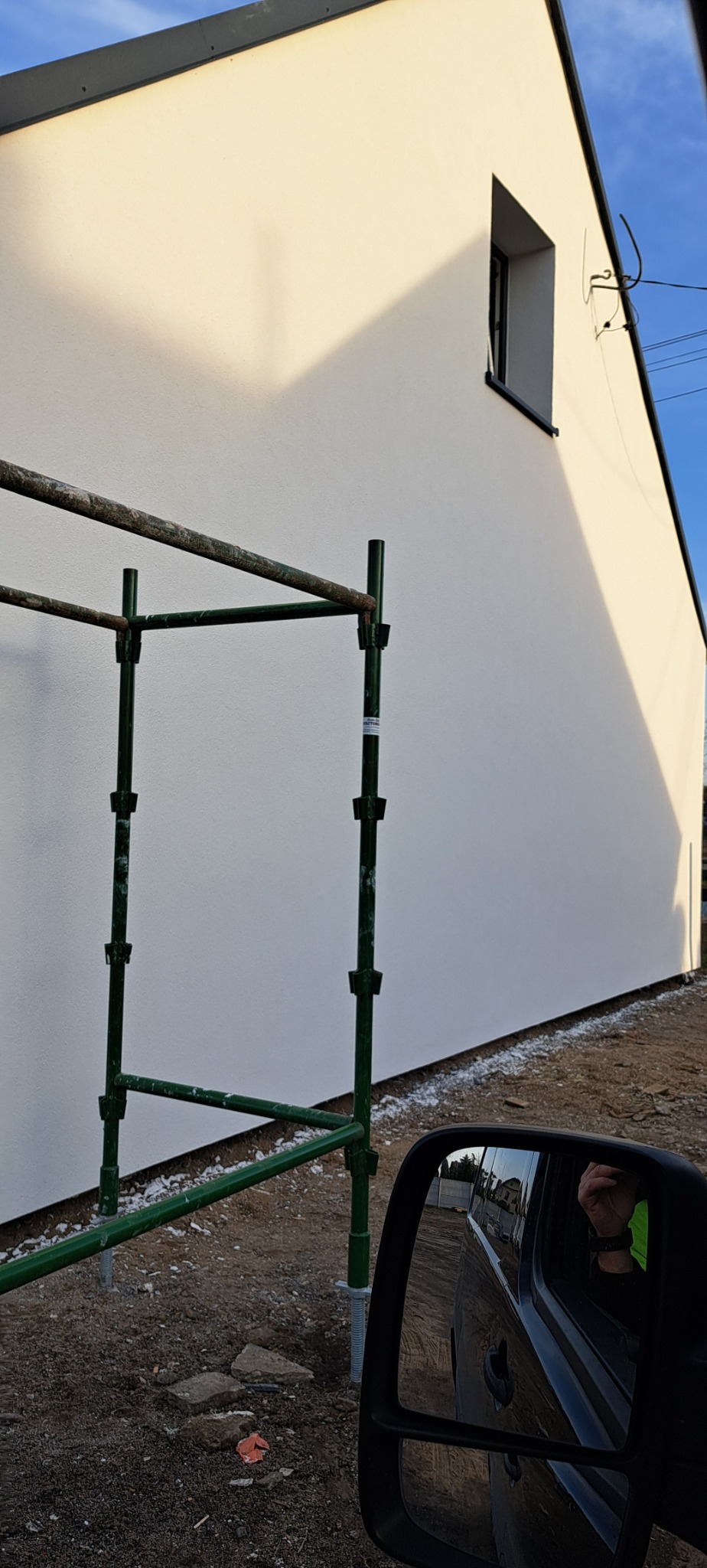 Świeżo pomalowana elewacja domu jednorodzinnego w jasnym odcieniu, widoczne rusztowanie z zielonych rur oraz odbicie w lusterku samochodu.