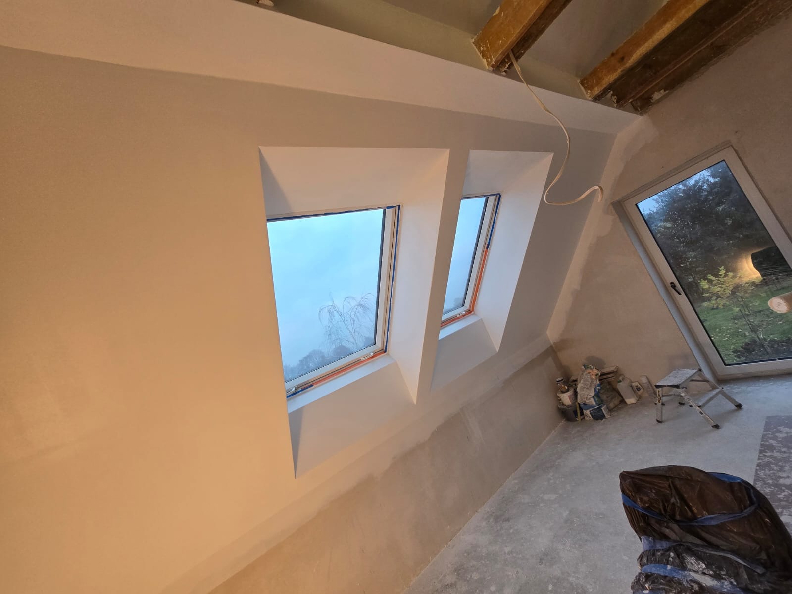 Poddasze w trakcie remontu: widoczne dwa okna dachowe, świeżo pomalowane ściany na biało, surowy beton na podłodze, drabina i materiały budowlane w rogu, oraz okno z widokiem na ogród.
