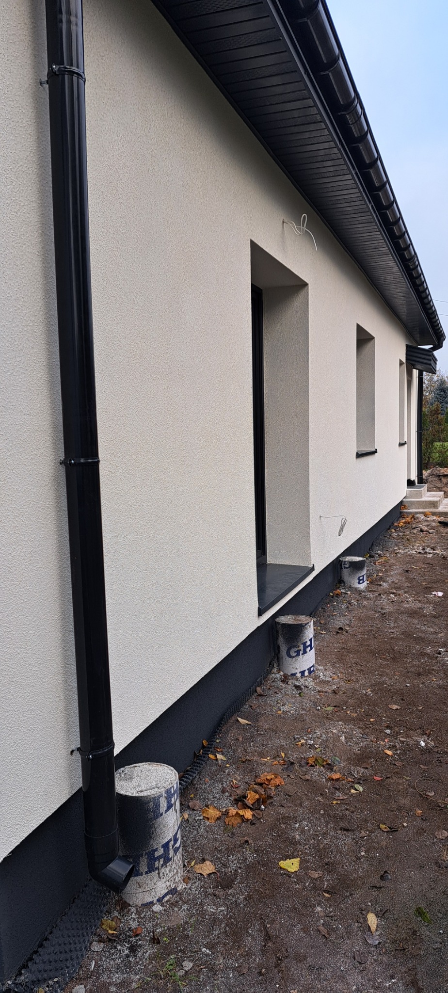 Fragment elewacji domu jednorodzinnego z czarną rynną, oknami z czarnymi ramami i wykończoną elewacją w kolorze złamanej bieli, widoczne zabezpieczenia wokół budynku.