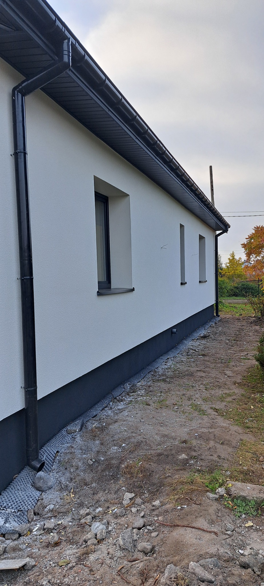 Świeżo otynkowana ściana domu jednorodzinnego z czarną rynną i podmurówką, widoczne trzy okna, teren wokół budynku jeszcze niezagospodarowany.