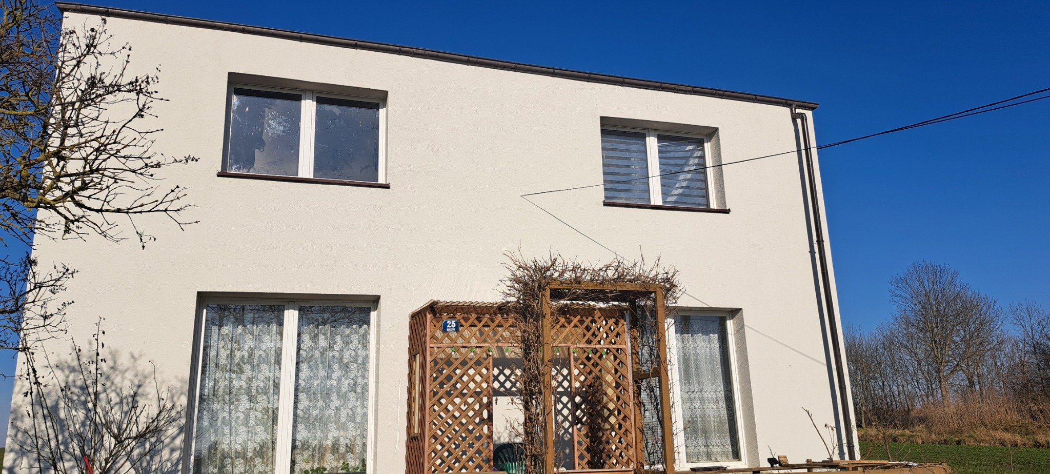 Dwukondygnacyjny dom z jasną elewacją, drewnianą kratką ozdobną przy wejściu i oknami z białymi ramami, widziany w słoneczny dzień.