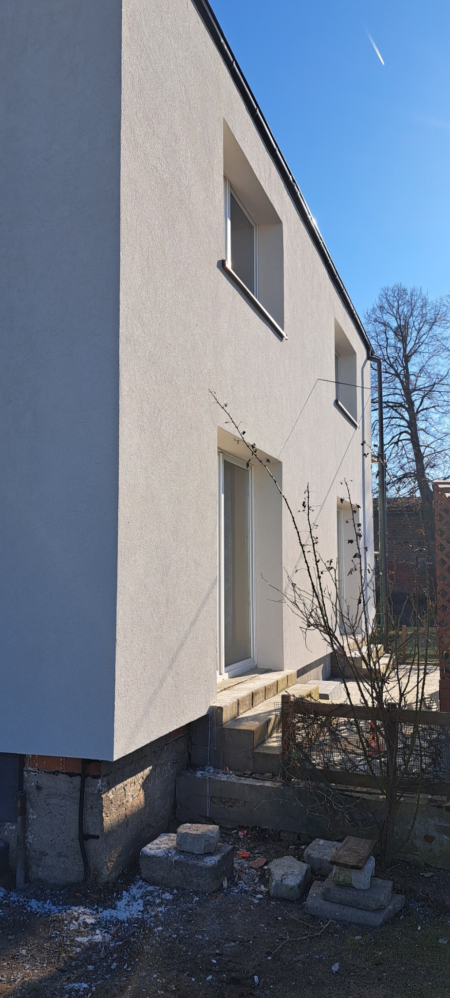 Wykończona elewacja domu jednorodzinnego z widocznym ociepleniem fundamentów, kamienne schody wejściowe i bezlistne krzewy na pierwszym planie, błękitne niebo w tle.