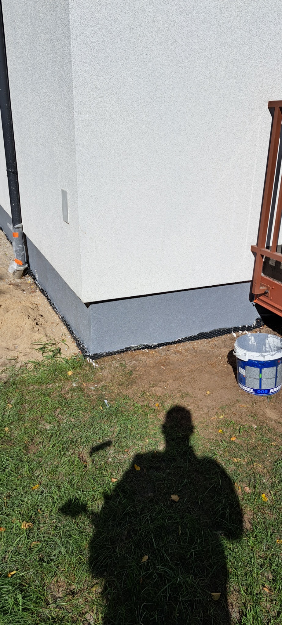 Dolna część elewacji budynku po malowaniu na szaro, widoczny kubeł z farbą i cień osoby malującej.