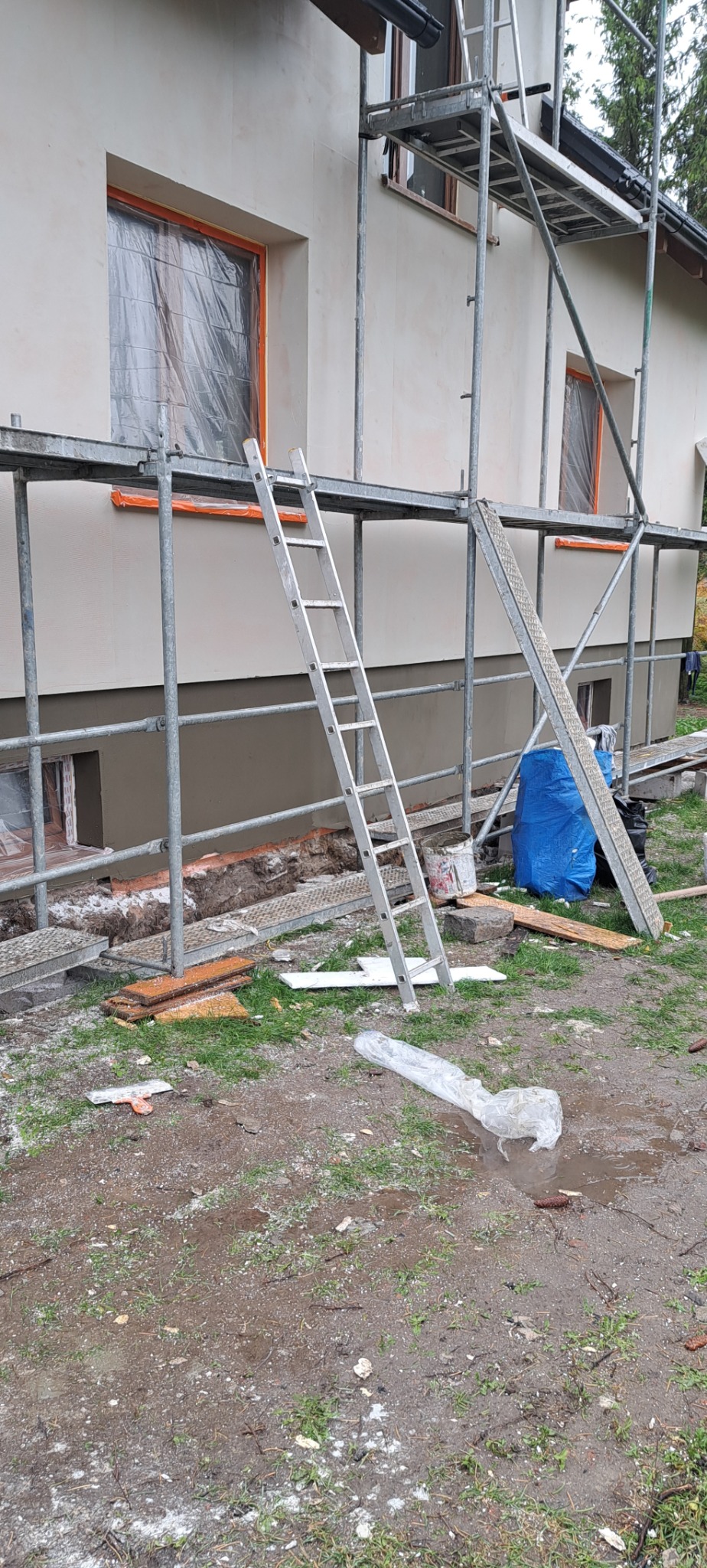 Dom jednorodzinny w trakcie prac elewacyjnych: ściany pokryte jasnym tynkiem, okna zabezpieczone folią, rusztowanie, drabina oparta o ścianę, widoczne materiały budowlane i narzędzia na ziemi.