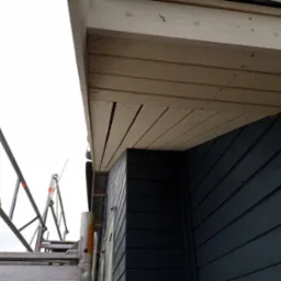 Fragment dachu z drewnianą podbitką w trakcie montażu, widok z rusztowania na elewację budynku z ciemnego drewna.