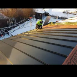 Pracownik w odblaskowej kamizelce montuje metalowe panele na dachu w zimowych warunkach; w tle ośnieżony krajobraz i rusztowanie.